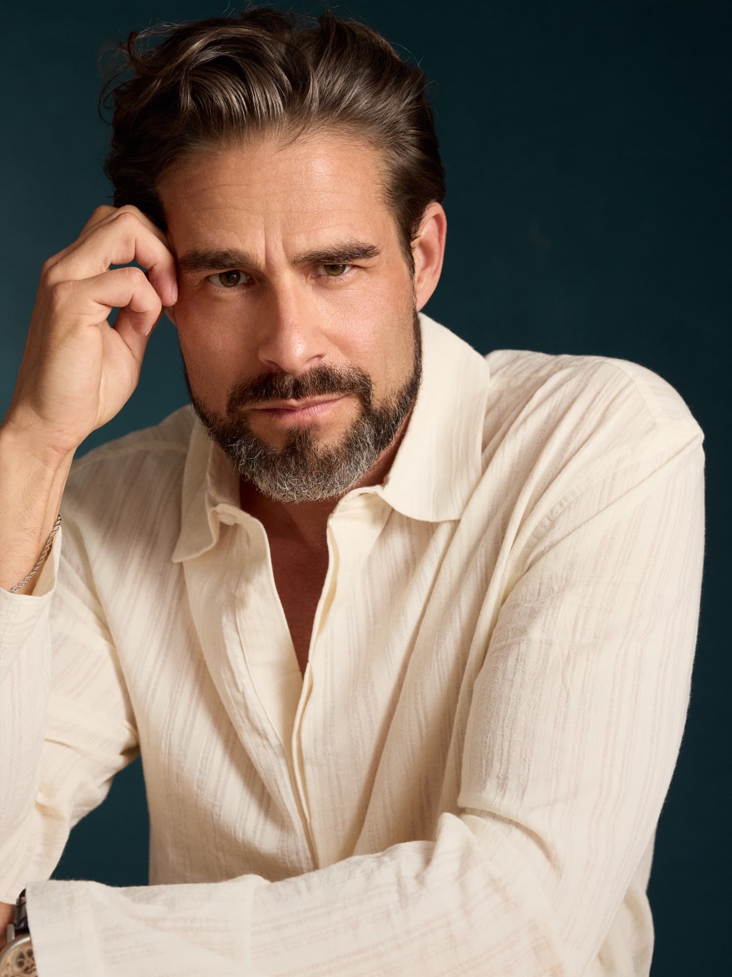 Brandon Mills — close editorial portrait in a textured cream shirt, hand to brow, teal backdrop
