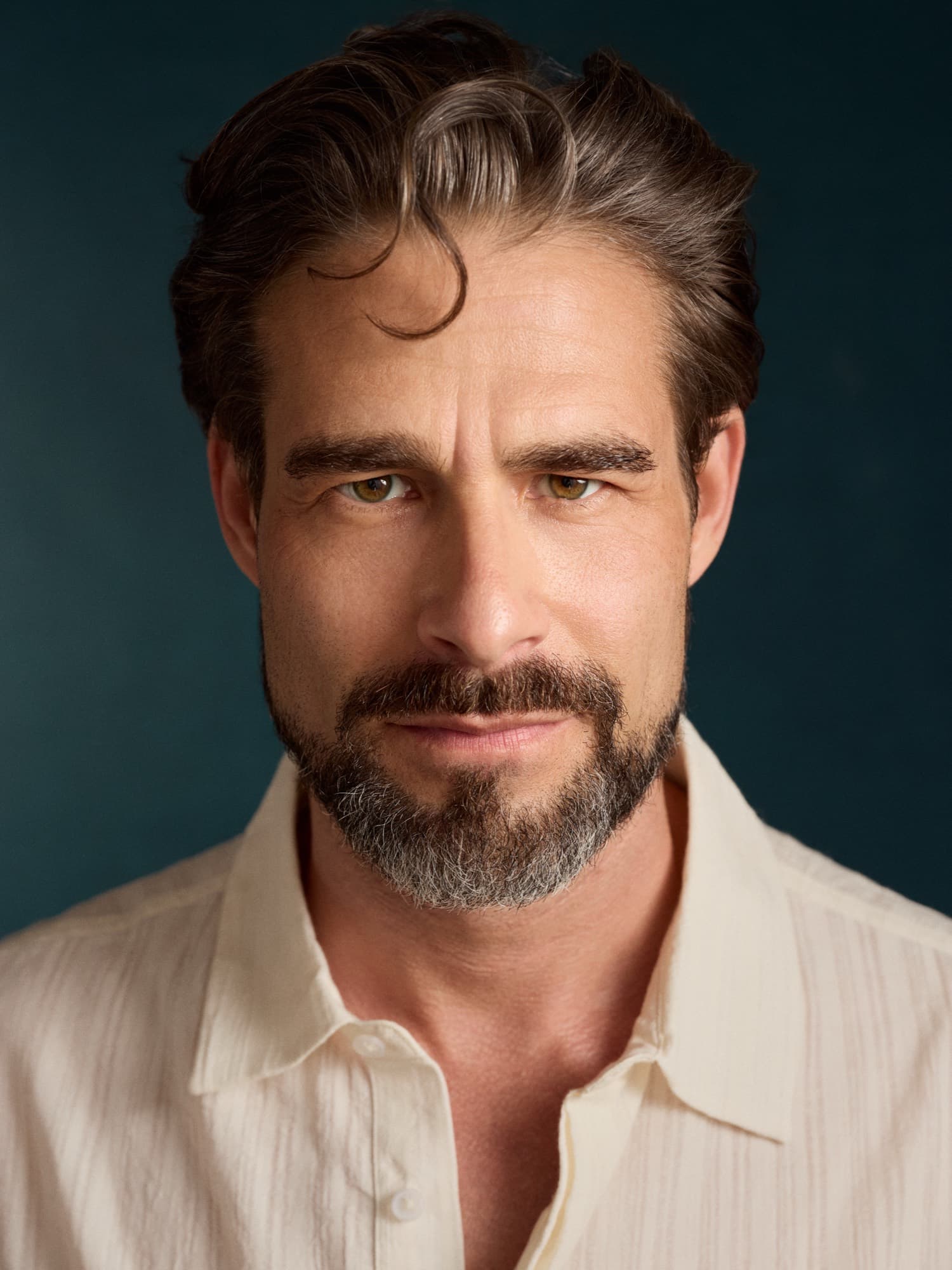 Brandon Mills — frontal close editorial headshot in a cream shirt against a teal backdrop
