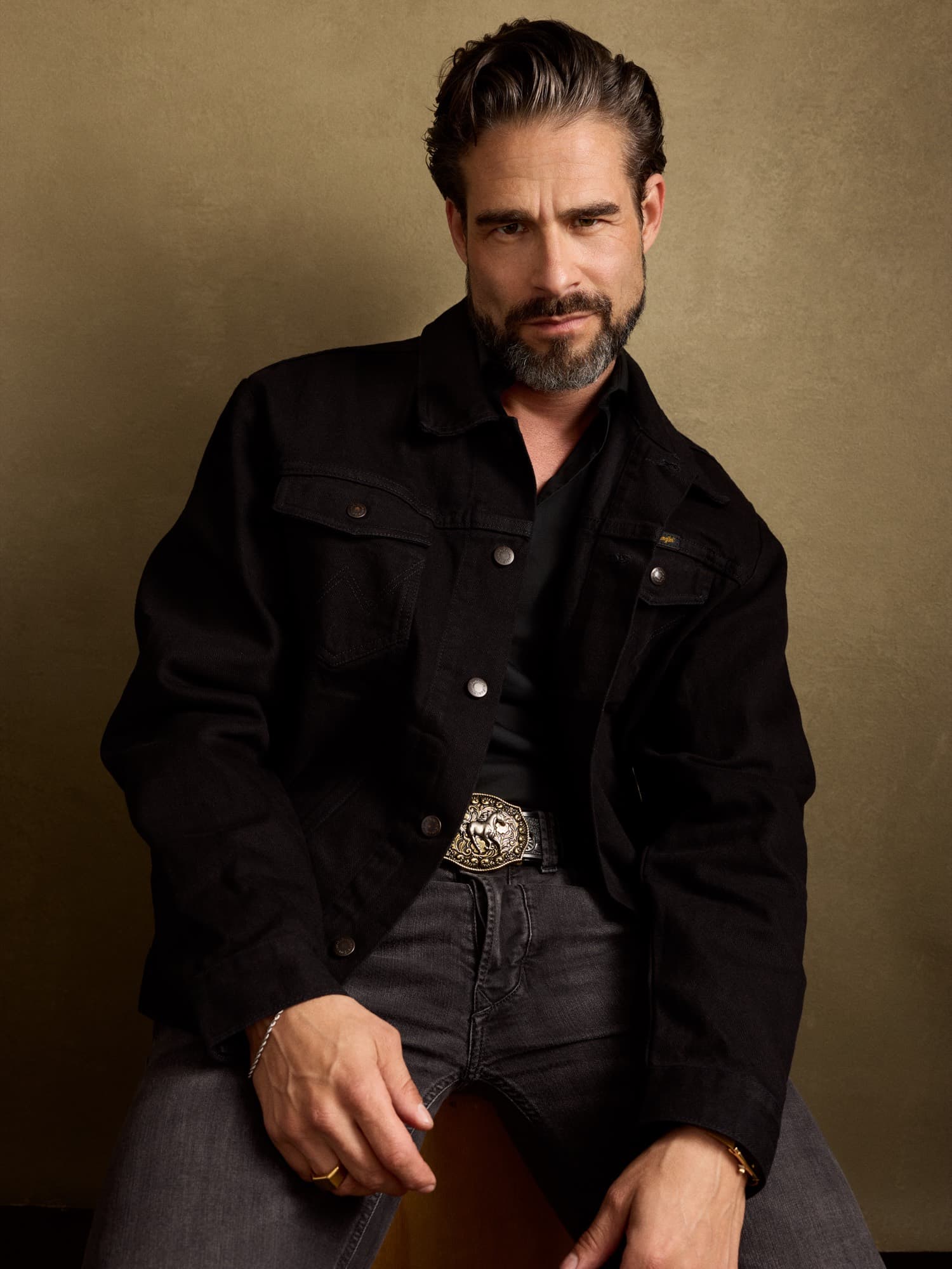 Brandon Mills — seated portrait in a black denim jacket with ornate belt against a warm painterly backdrop